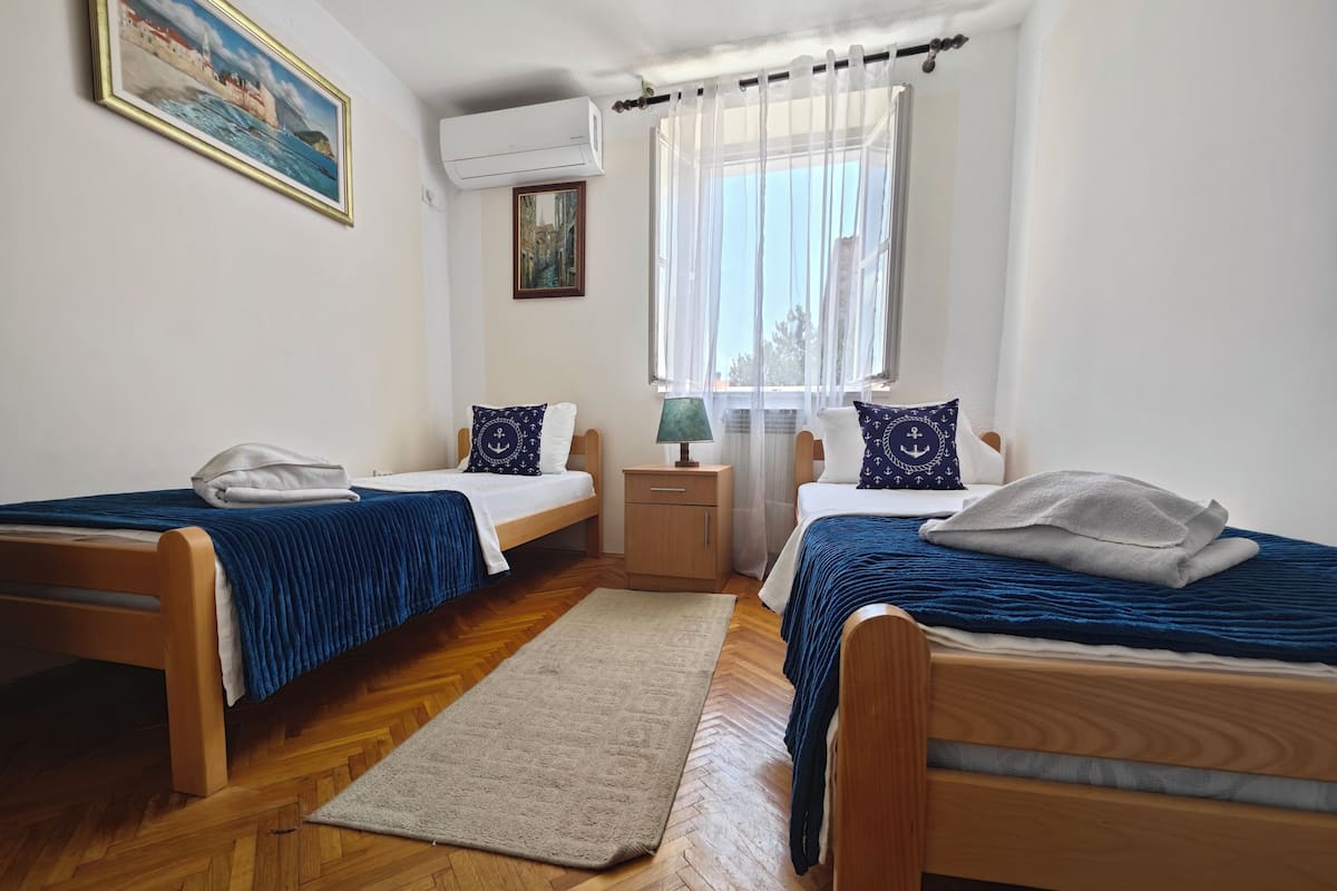 A room with two wooden beds, a small bedside table with a lamp, a wooden floor, and a window with sheer curtains.