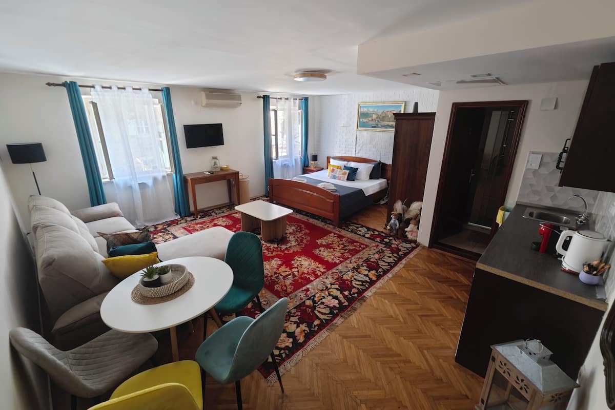 A compact hotel room with a bed, a small desk, a sofa, a dining table, and a kitchen area.