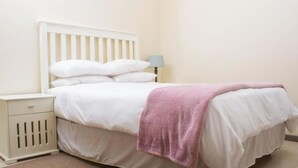 Quarto individual com uma cama de solteiro, cabeceira branca, mesa de cabeceira com abajur e manta rosa.