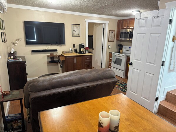 A living room with a brown leather couch, a wooden dining table, a flat-screen TV, a kitchen area with a microwave and oven, and a door leading to another room.