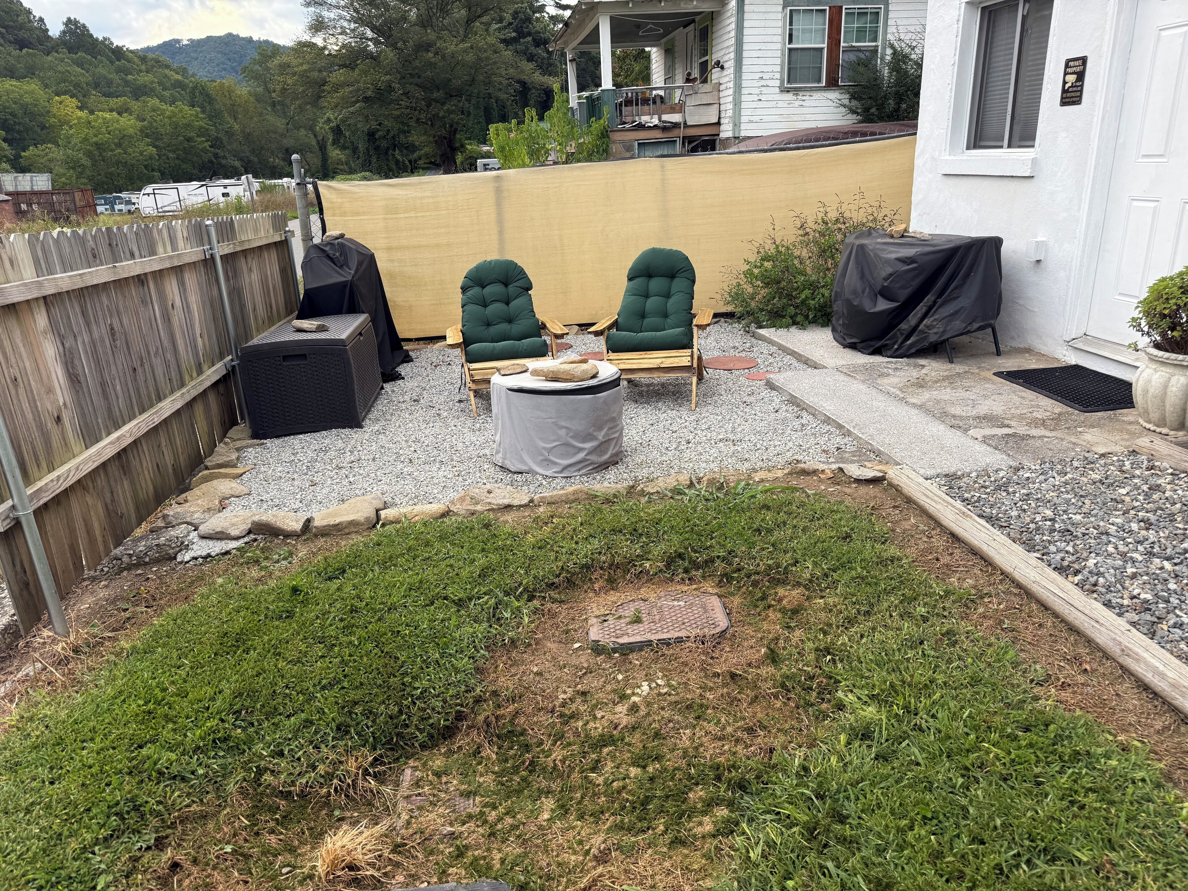 A backyard with a wooden fence, two green chairs, a covered grill, and a small table.