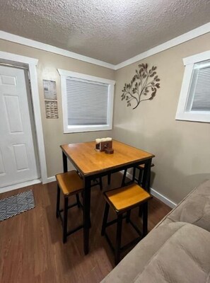 A compact living space with a dining table and chairs, a sofa, a decorative wall piece, and a window with blinds.