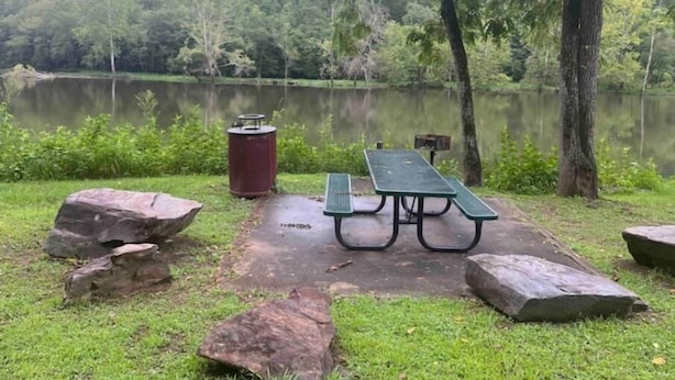 Una mesa de picnic con bancos, un basurero y rocas grandes junto a un lago en un parque.
