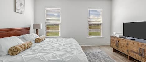 A bedroom with a bed, a television, a wooden cabinet, and two windows with blinds.