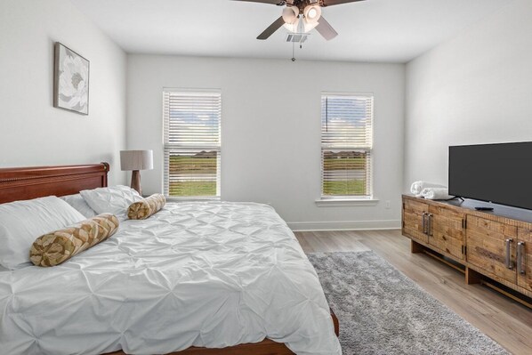 A bedroom with a bed, a television, a wooden cabinet, and two windows with blinds.