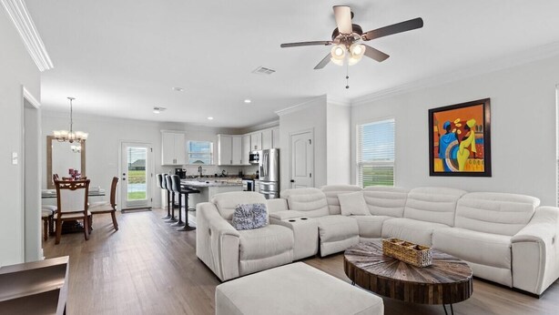 A modern living room with a sectional sofa, a dining area with chairs, a kitchen with appliances, and a ceiling fan.