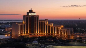 A large hotel with a prominent tower and multiple columns, surrounded by a parking area and distant cityscape.