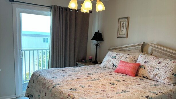 A bedroom with a large bed, a ceiling fan, a nightstand, a lamp, and a view of the outside balcony.