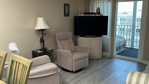 A living room with a flat-screen TV, a ceiling fan, a sofa, a dining table with chairs, and a view of the outside balcony.