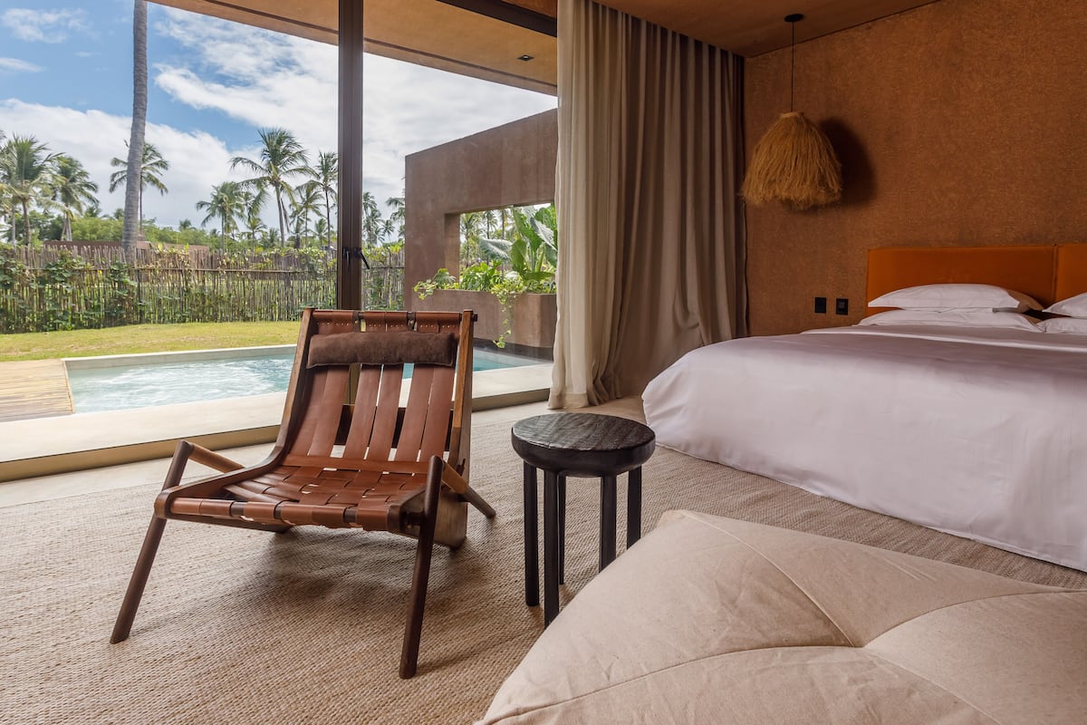 Quarto com uma cama grande, uma cadeira de madeira, uma mesa redonda pequena e vista para uma piscina e palmeiras.