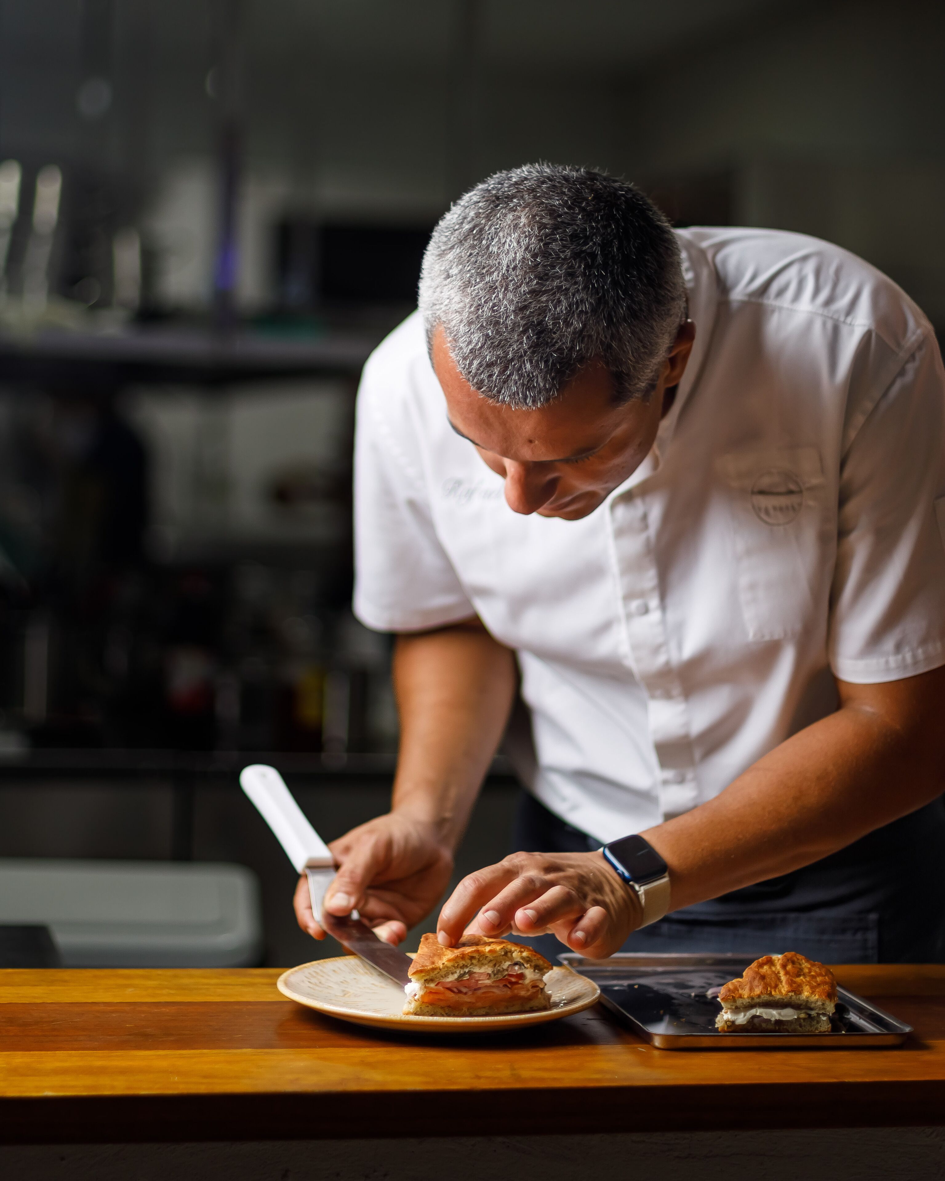 A chef slicing a sandwich on a wooden table in a kitchen.