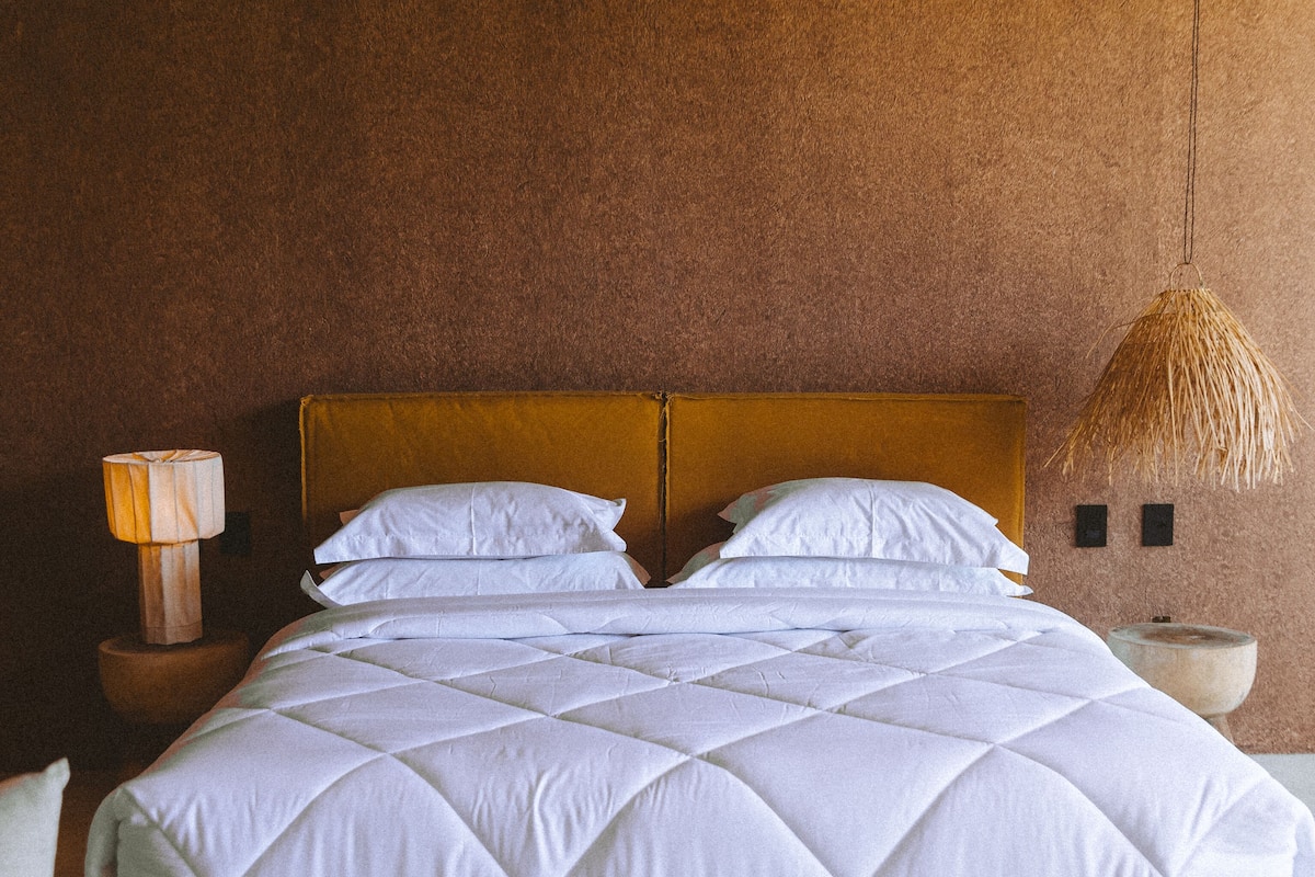 Cama com estrutura de madeira, roupa de cama branca e cabeceira. Apresenta uma mesa de cabeceira com abajur e um relógio fixado na parede.