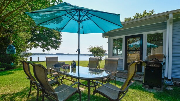 An outdoor patio with a glass table and chairs, a blue umbrella, and a view of a lake.
