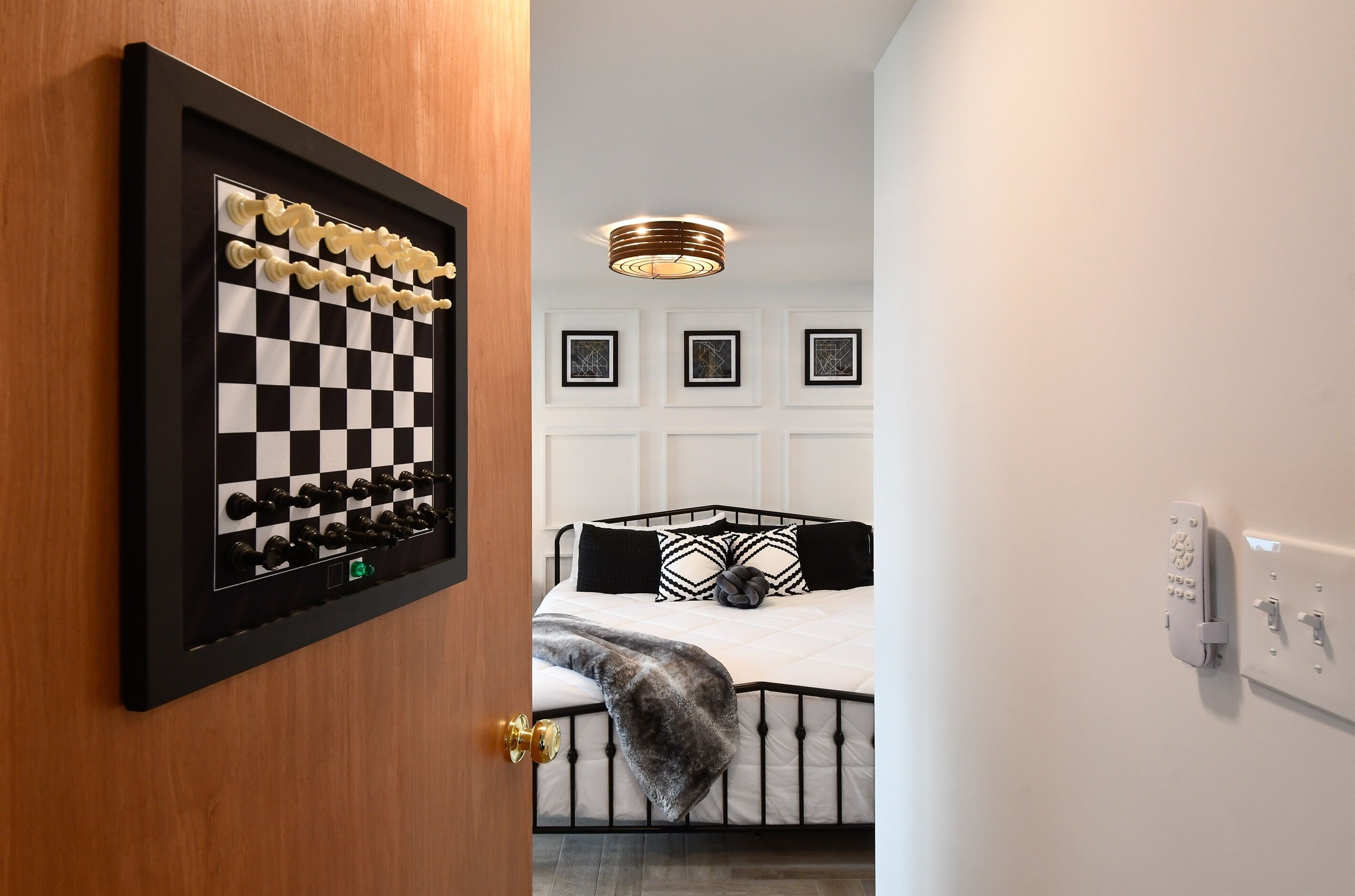 A bedroom with a chessboard, a bed with black and white bedding, a ceiling light, and framed artwork on the wall.