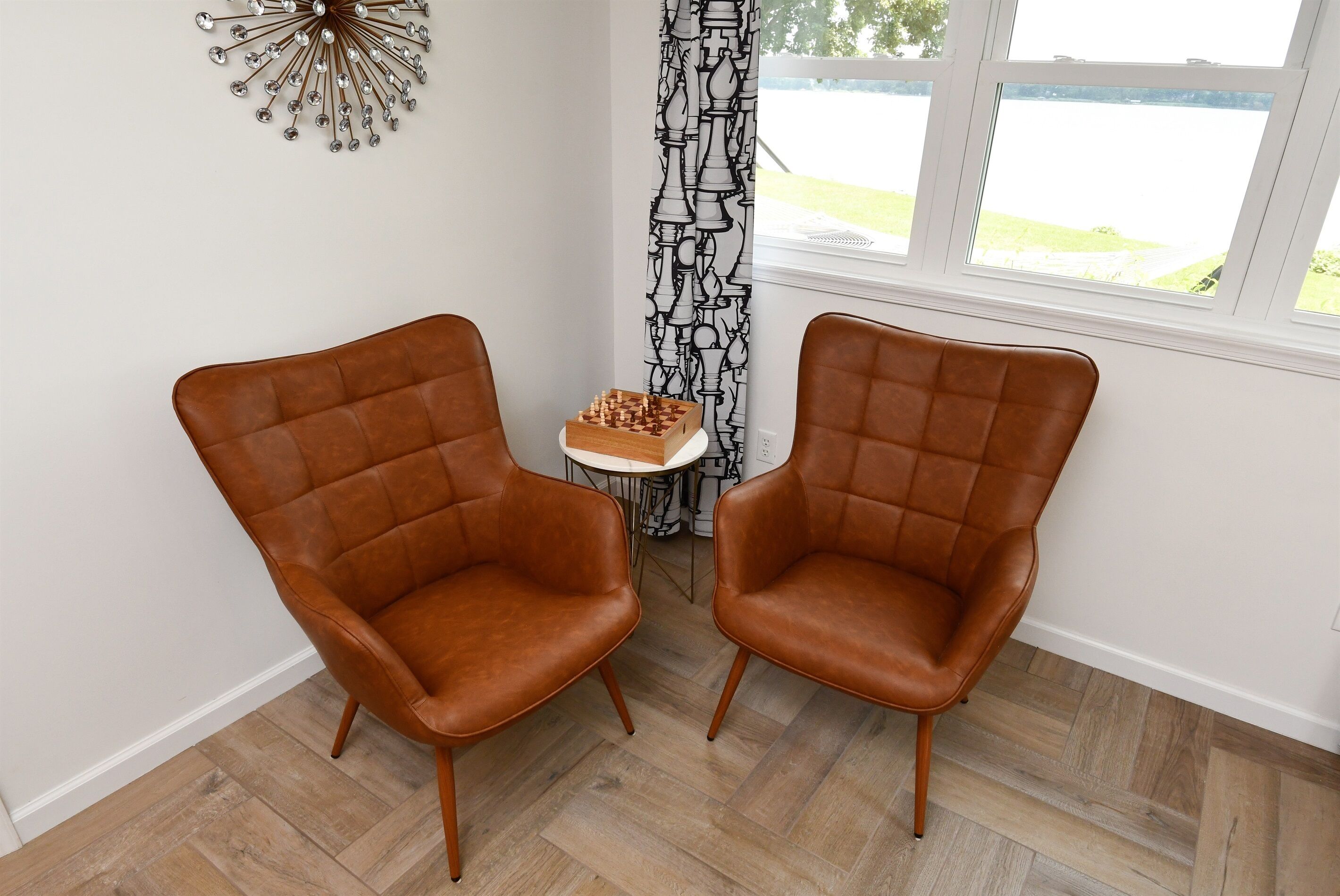 A room with two brown leather chairs facing a small table with a chessboard. The room has a window with a view of a body of water and a patterned curtain.