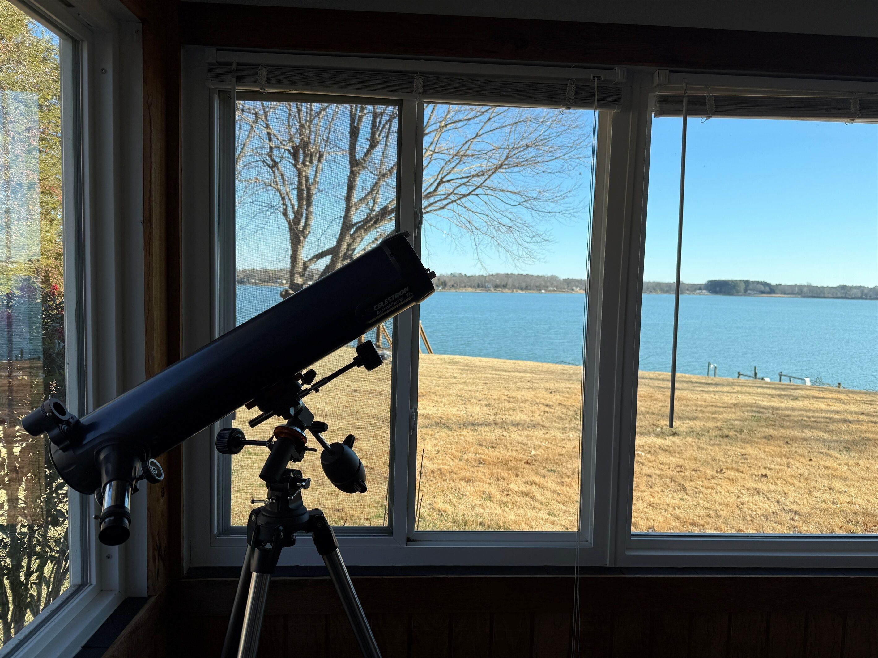 A telescope on a tripod inside a room with large windows overlooking a lake.