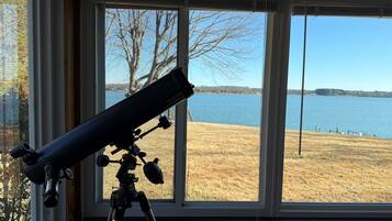 A telescope on a tripod inside a room with large windows overlooking a lake.