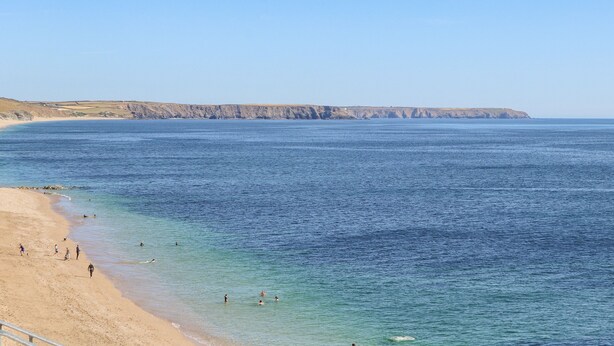 Uma praia costeira com águas azuis cristalinas, areal arenoso e falésias rochosas ao fundo.