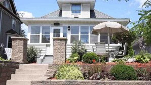 Ein zweistöckiges Haus mit Veranda, einem Steinpfeiler und einem kleinen Garten mit unterschiedlichen Pflanzen und einem Patio-Tisch.