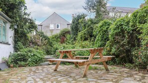 Una mesa de picnic de madera en un jardín con pavimento de piedra, rodeada de abundante vegetación y una casa al fondo.