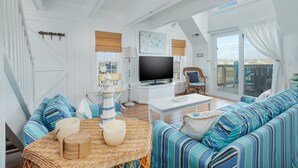 A spacious living room with a large TV, striped sofas, a wicker table, and a view of the outside. - 2910 - Dolphin's Den, Duck Condo | Pools, Tennis & Views (Duck)