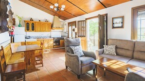 A cozy living room with a sofa, armchair, and wooden dining table. A kitchen with wooden cabinets and a stove is visible in the background. - Cubo's Casa Rural Jose & Caminito del Rey / Piscina privada / Caminito del Rey (Ardales)
