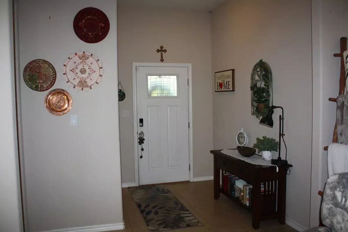A hallway with a white door, a welcome sign, a mirror, and a small table with a lamp.