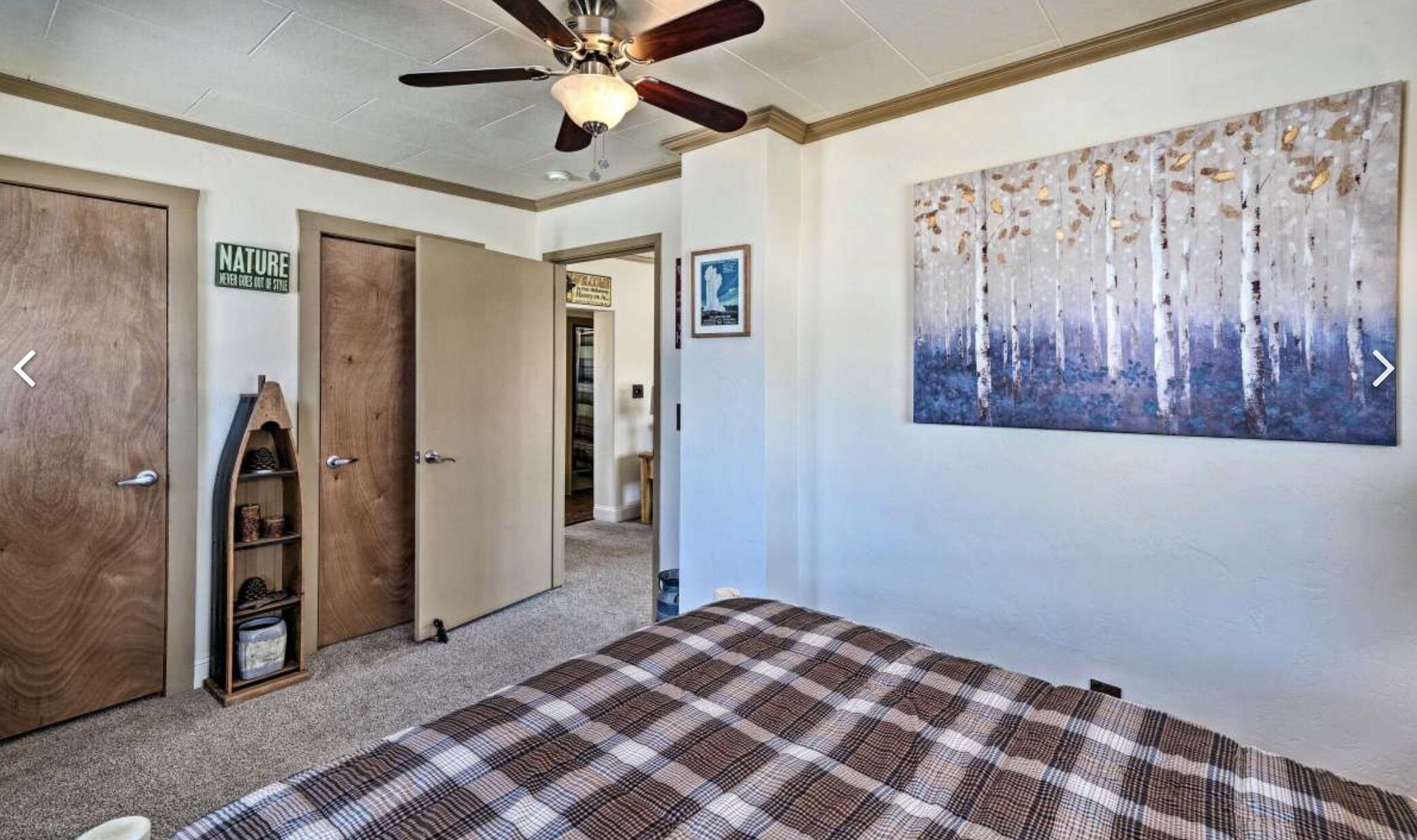 A bedroom with a ceiling fan, a large painting of birch trees, a plaid bedspread, and a wooden door with a 'Nature' sign.