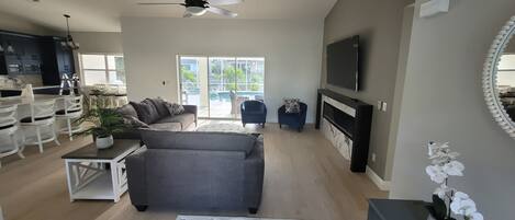 A modern living room with a grey sofa, a dining area with white chairs, a TV mounted on the wall, and a large window offering a view of the outdoors.