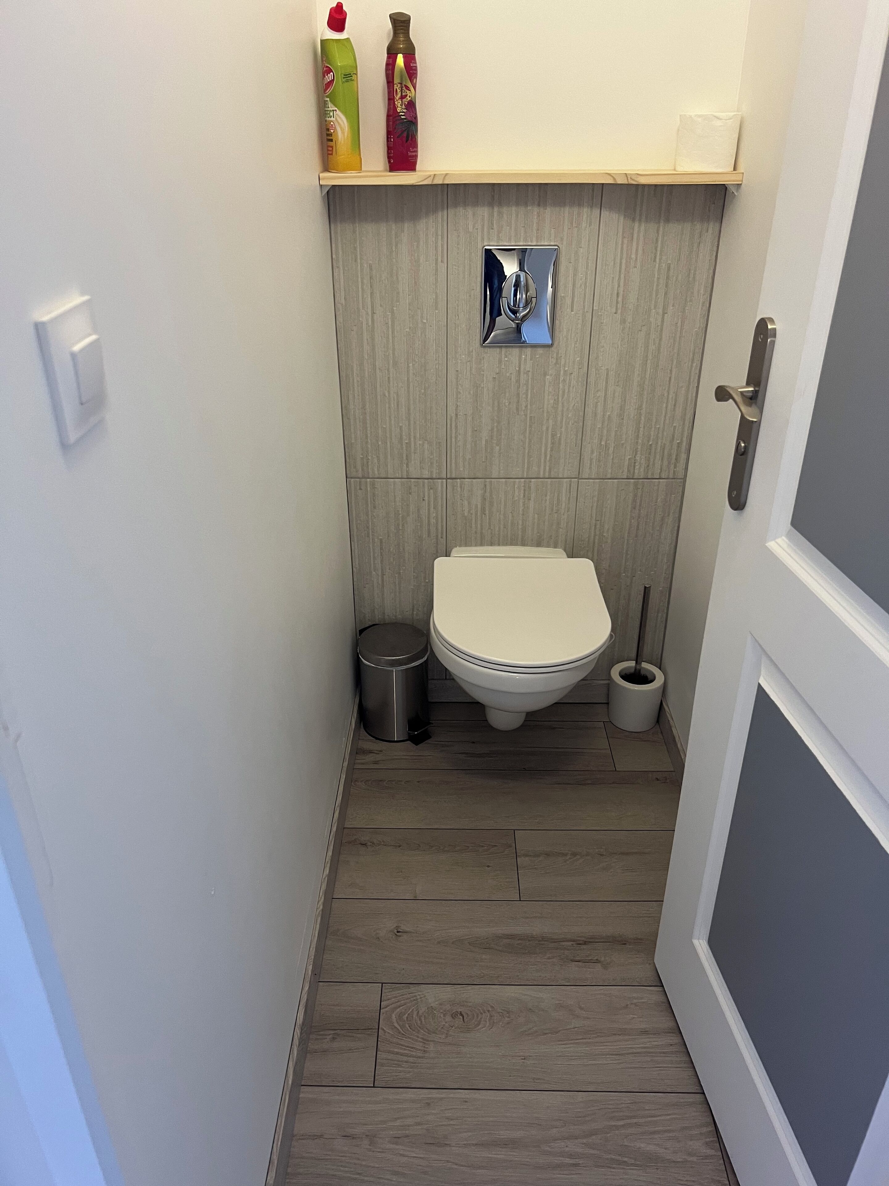 Un baño pequeño con inodoro, un cubo de basura y un bidé. Hay un estante sobre el inodoro con dos botellas.