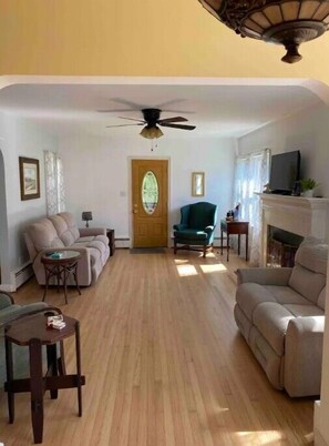 A spacious living room with a wooden floor, a ceiling fan, a fireplace, and a television.