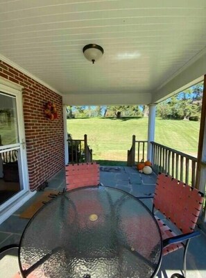 A covered porch with a glass table and chairs, a brick wall, and a view of a grassy yard.