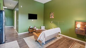 A room with a green wall, a sofa, a TV, a wooden cabinet, and a floor lamp. - Quiet End-Unit Studio w/ Full Kitchen + Natural Light (Portland)