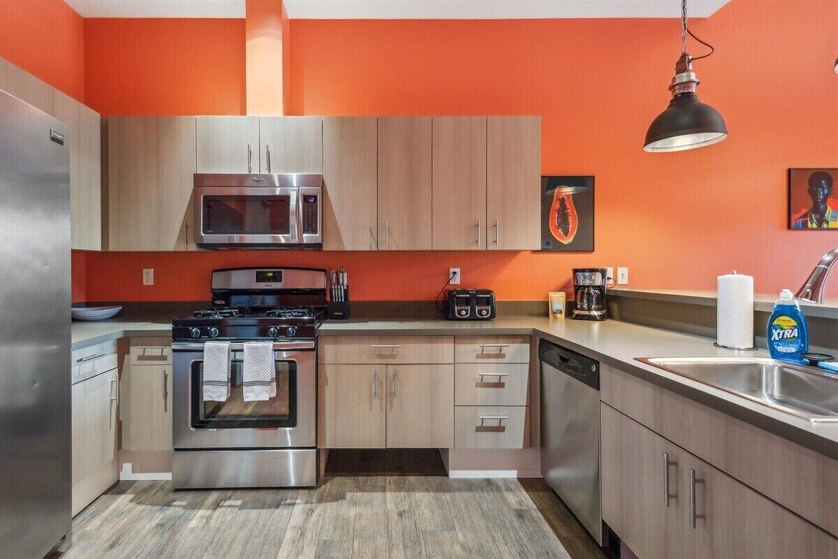 A modern kitchen with stainless steel appliances, wooden cabinets, and a bright orange wall.