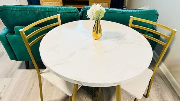 Een ronde eettafel met een witte marmeren blad, omringd door vier stoelen met gouden poten. Op de tafel staat een vaas met witte bloemen.