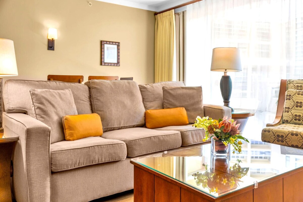 Une chambre d’hôtel avec un canapé beige, une table basse en bois, un fauteuil à motifs et un vase rempli de fleurs.