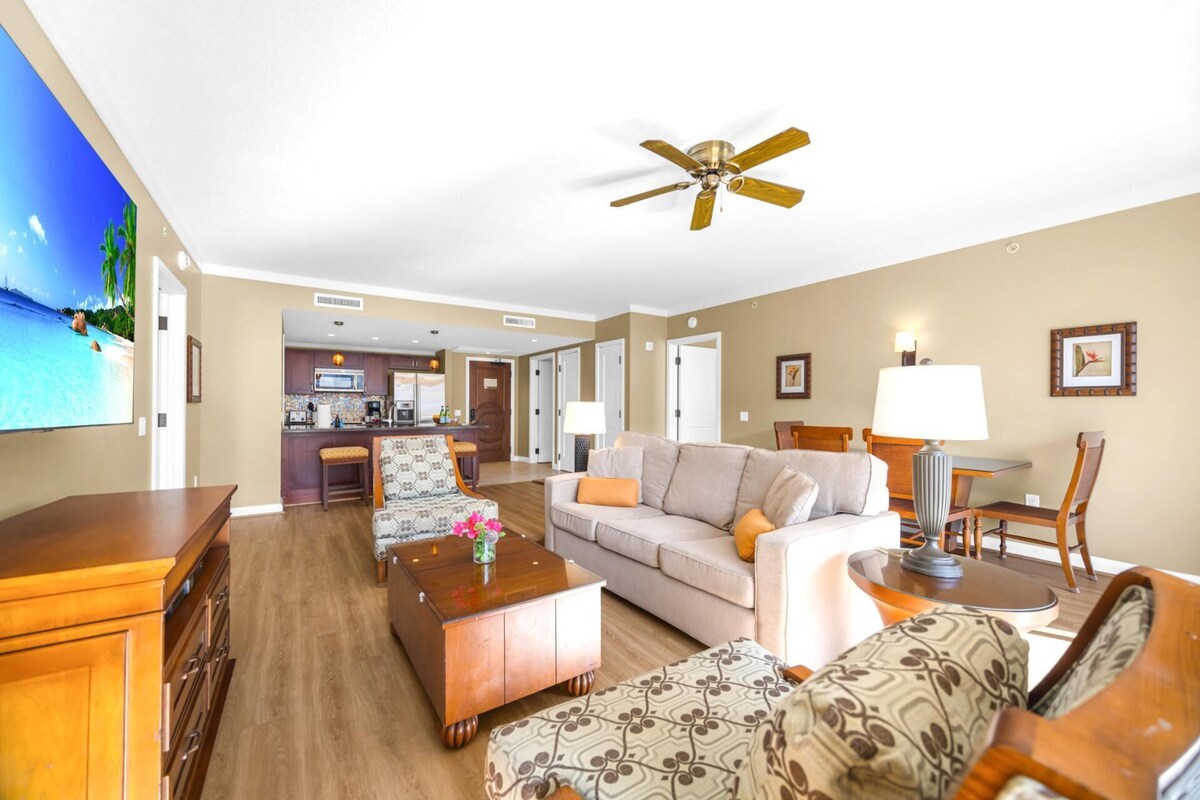 A hotel room with a living area, a kitchenette, a ceiling fan, and a TV displaying a tropical beach scene.