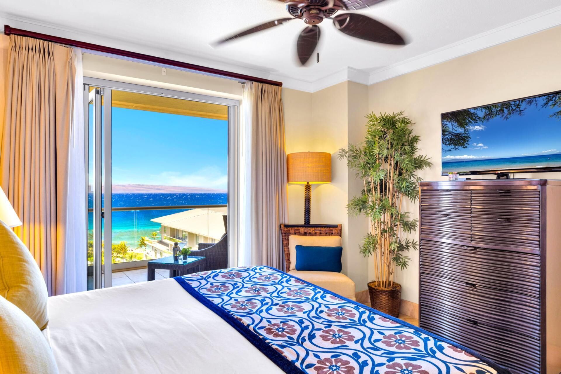 Ein Hotelzimmer mit Bett, Fernseher, Deckenventilator, Fenster mit Meerblick und einer Pflanze.