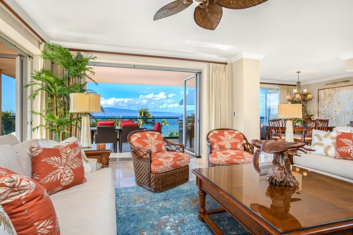 A spacious living room with a large glass door leading to a balcony with a view of the ocean. There are comfortable seating areas with patterned cushions and a dining area with a chandelier.