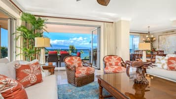 Un vaste salon doté d’une grande porte vitrée donnant sur un balcon avec vue sur l’océan. On y trouve des coins salon confortables avec des coussins à motifs, ainsi qu’un coin repas agrémenté d’un lustre.
