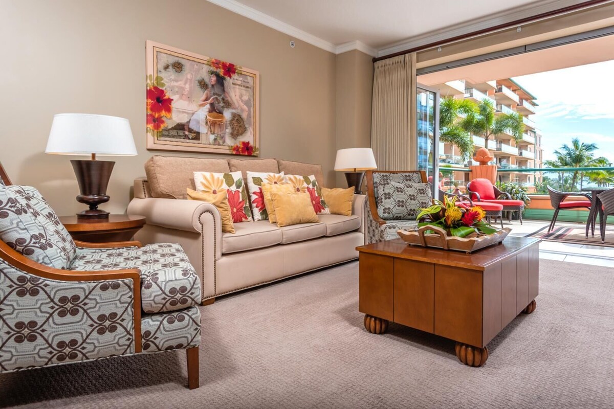 Une chambre d’hôtel avec un canapé beige, des fauteuils à motifs, une table basse en bois et une grande fenêtre donnant sur un palmier et un immeuble.