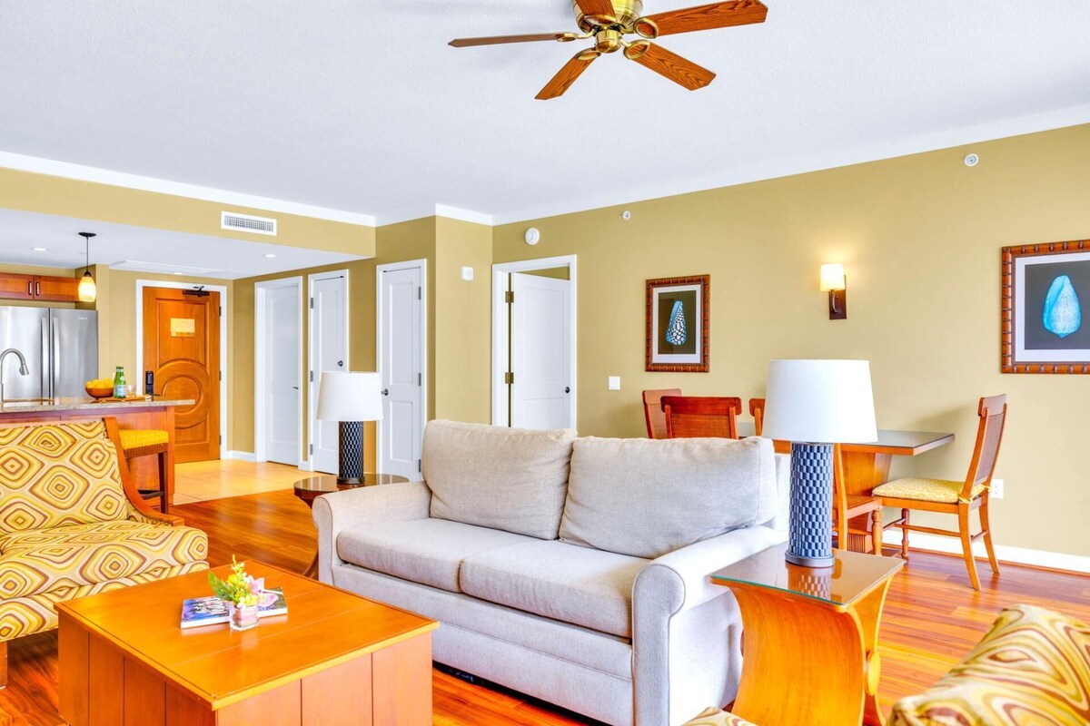 A spacious living room with a sofa, coffee table, and chairs. There is a kitchen area with a refrigerator and a dining table with chairs. The room has wooden flooring and a ceiling fan.