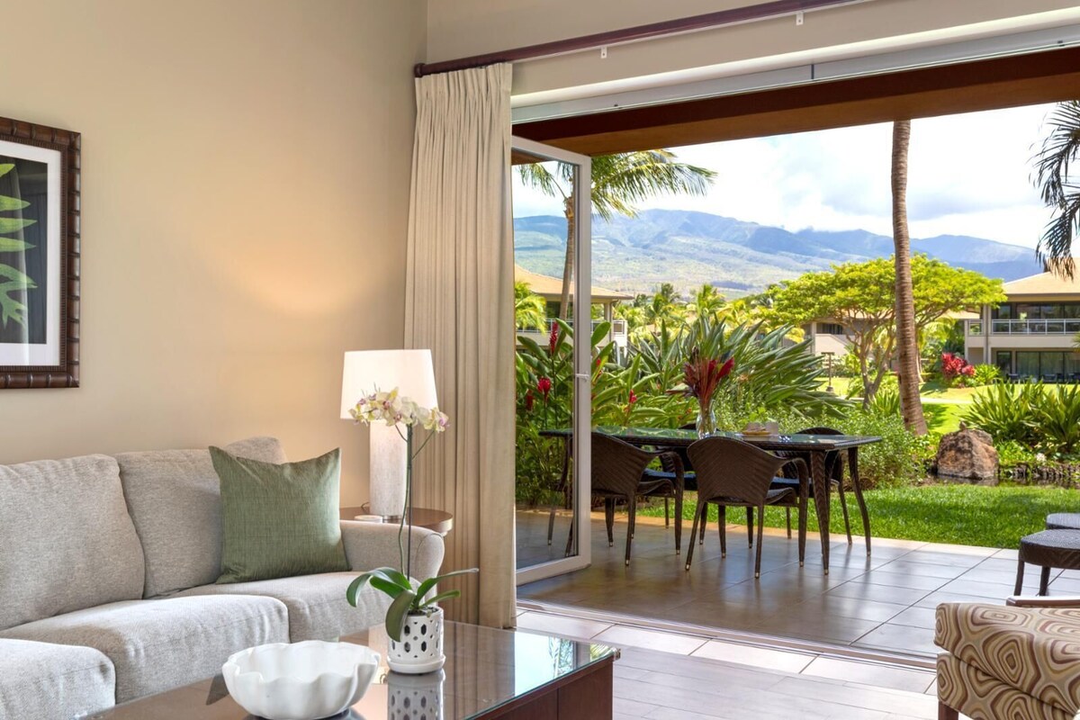 Une chambre d’hôtel avec un canapé, une table avec une lampe et une vue sur un jardin luxuriant et des montagnes.