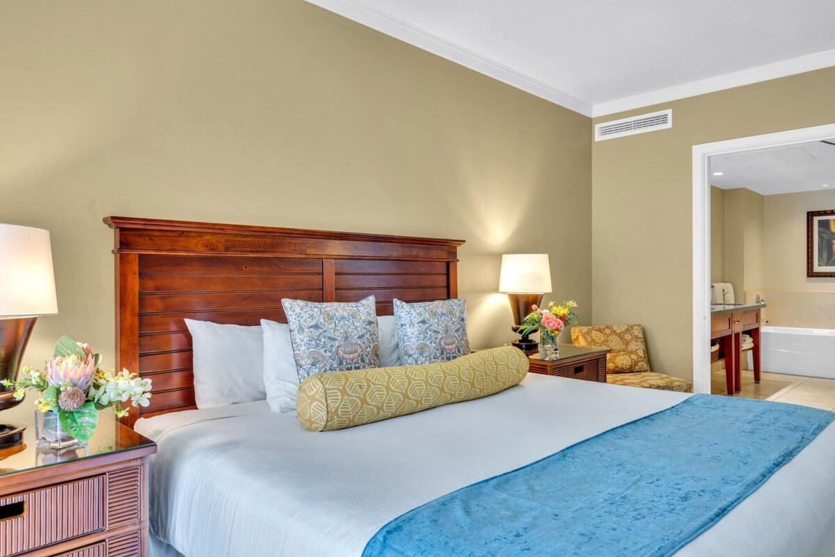 Una habitación de hotel con cabecera de madera, una cama grande con ropa de cama estampada, una mesita de noche con lámpara y una pequeña mesa con un jarrón de flores.