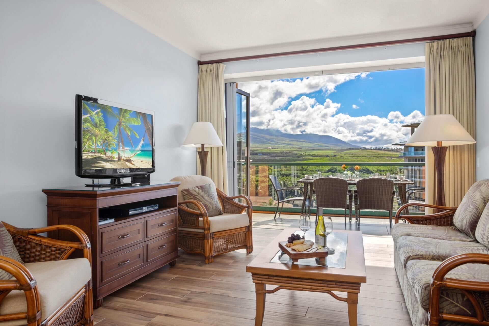 A spacious living room with a large flat-screen TV, wicker furniture, and a balcony with a scenic view.
