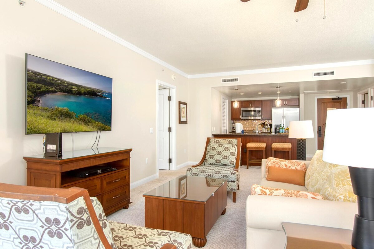 A living room with a flat-screen TV, a wooden entertainment center, patterned armchairs, a sofa, and a glass coffee table.