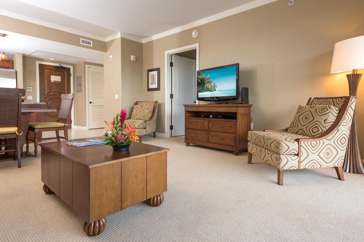 Une chambre d’hôtel avec une table basse en bois, un fauteuil à motifs, un téléviseur posé sur un meuble en bois et un coin repas avec une table et des chaises en bois.