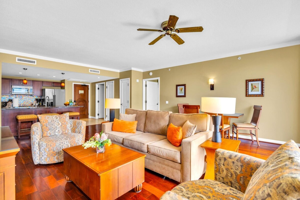 A spacious living room with a beige sofa, patterned armchairs, a wooden coffee table, and a ceiling fan. A kitchen with dark wooden cabinets and a tiled backsplash is visible in the background.
