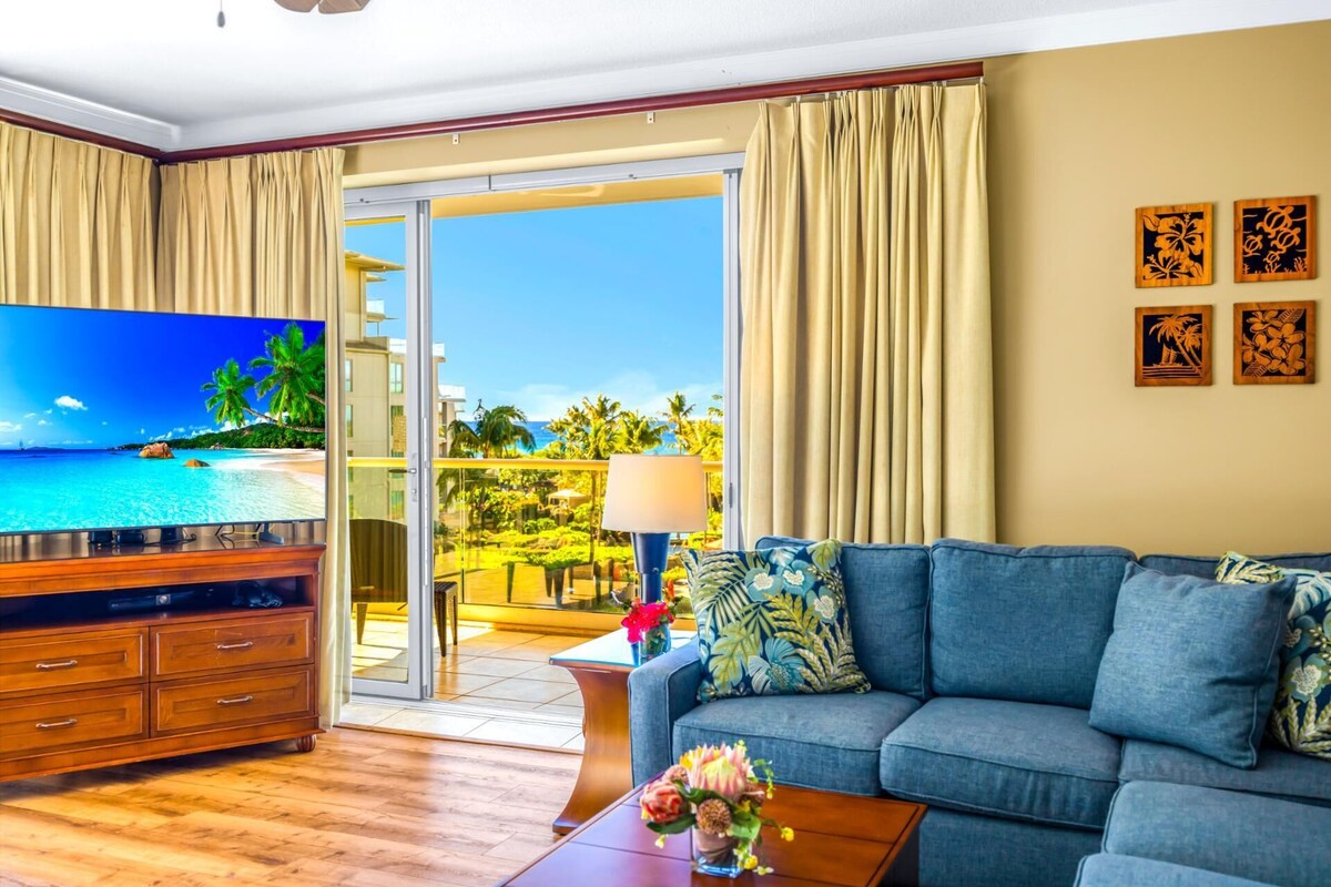 A living room with a large TV, a sofa, a coffee table, and a view of a beach through a sliding glass door.