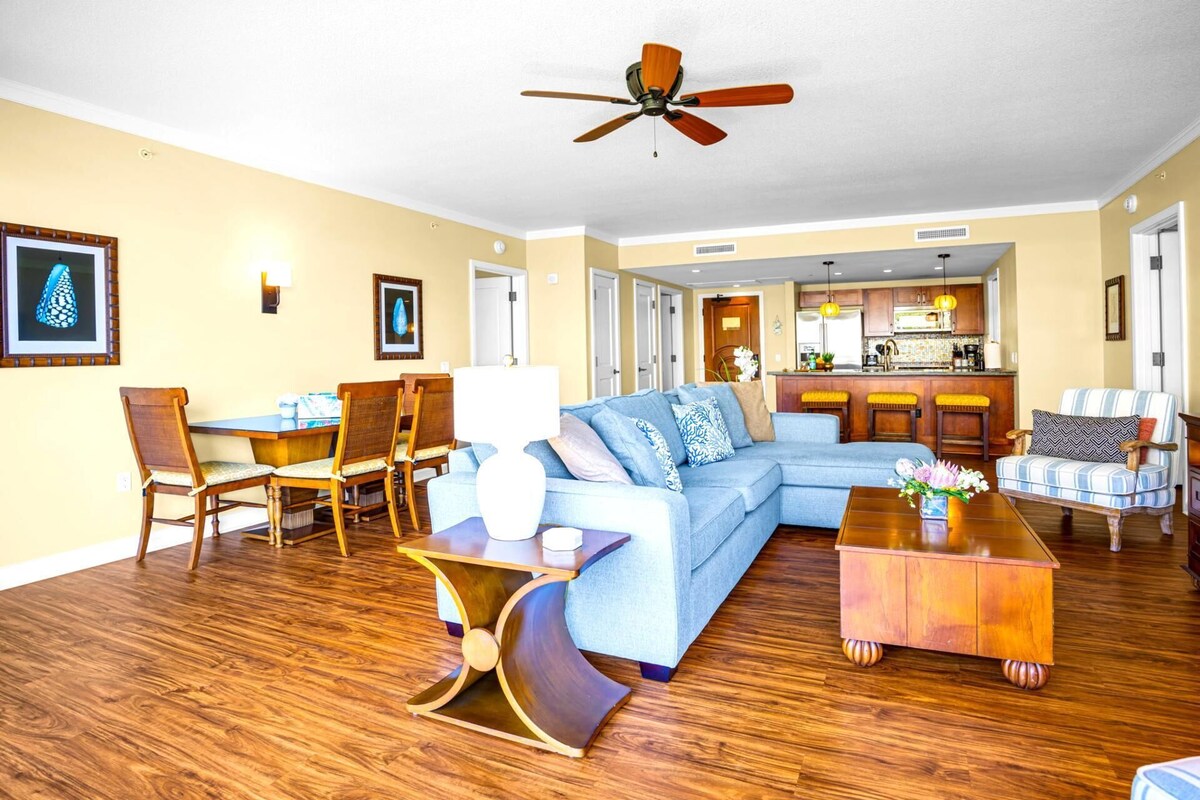 Un vaste salon comprenant un canapé d’angle, une table basse en bois et un coin repas avec des chaises. On aperçoit également un ventilateur de plafond et une cuisine à l’arrière-plan.